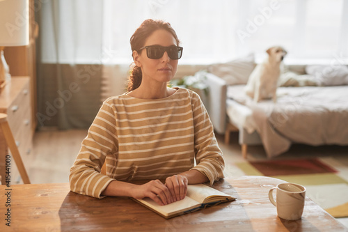 Warm toned portrait of modern blind woman reading braille book while sitting at table in cozy home interior with guide dog in background, copy space