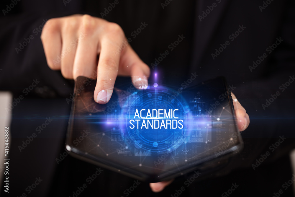 Naklejka premium Young man holding a foldable smartphone with ACADEMIC STANDARDS inscription, educational concept