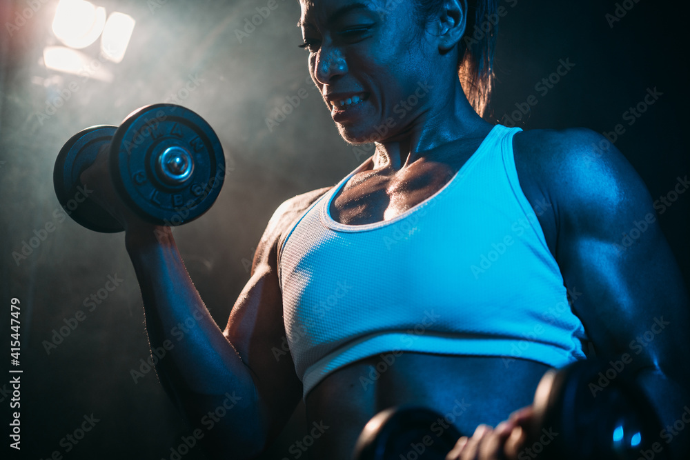 Close up shot of body builder athletes lifting dumbbell with dark tone lighting and fog atmosphere in the background. Selective focused. Self challenge, Muscular woman. Concept tough to  be stronger.