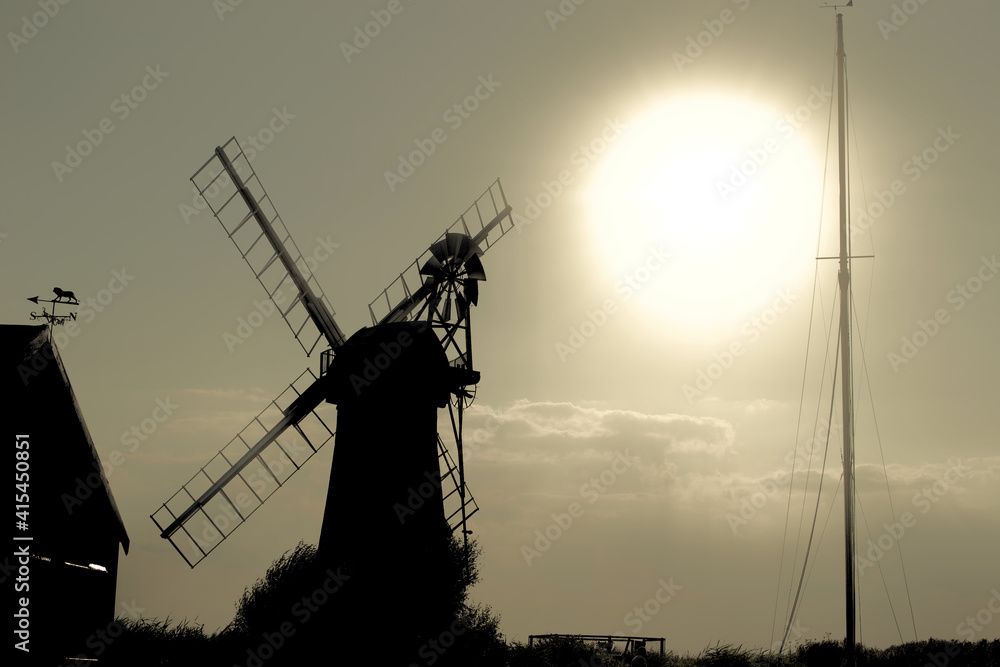 A traditional windmill (wind pump) silhouetted against a large bright low level evening sun. Shadows and darkness shroud the traditional building and a sailing boat's mast in the Norfolk Broads, UK