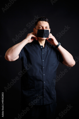 A young male doctor in a black surgical suit puts on a black mask to protect against the virus. Isolated on black background © Дмитрий Ткачук