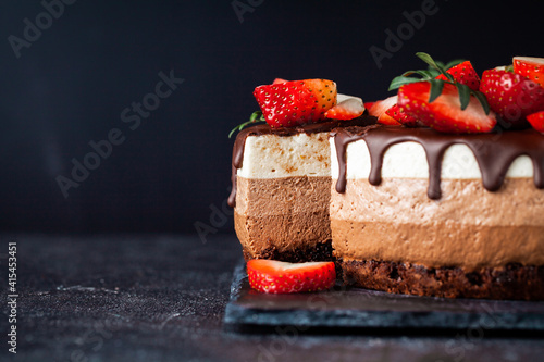 Three chocolates cake with chocolate drips on a black background. Layered cake with milk, black and white chocolate souffle decorated with strawberries on top. Confectionery background with copy space photo