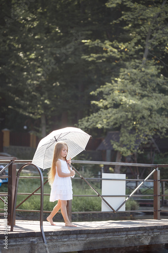A beautiful girl of six years old in a short white dress stands on the masonry near the river and holds a white umbrella in her hands
