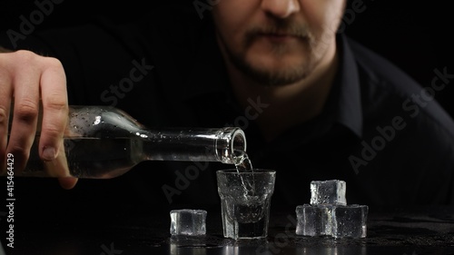 Bartender man pouring up frozen vodka or sake from bottle into shot glass with ice cubes on black background. Barman pour of clear transparent alcohol drink rum or tequila in shot-glass and drinking