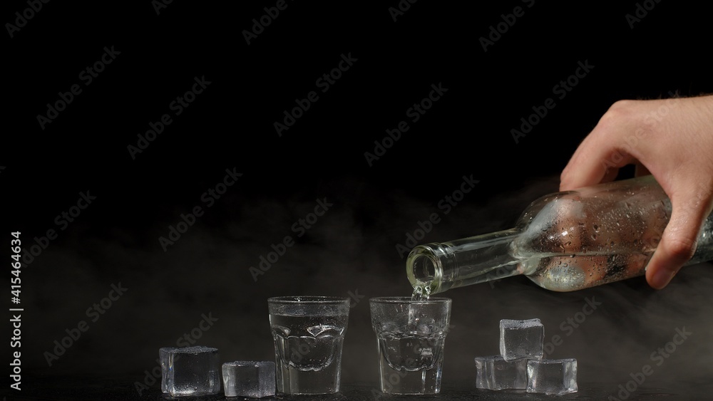 Αυτοκόλλητο Bartender pouring up two shots of vodka with ice cubes from a bottle into glasses against black background