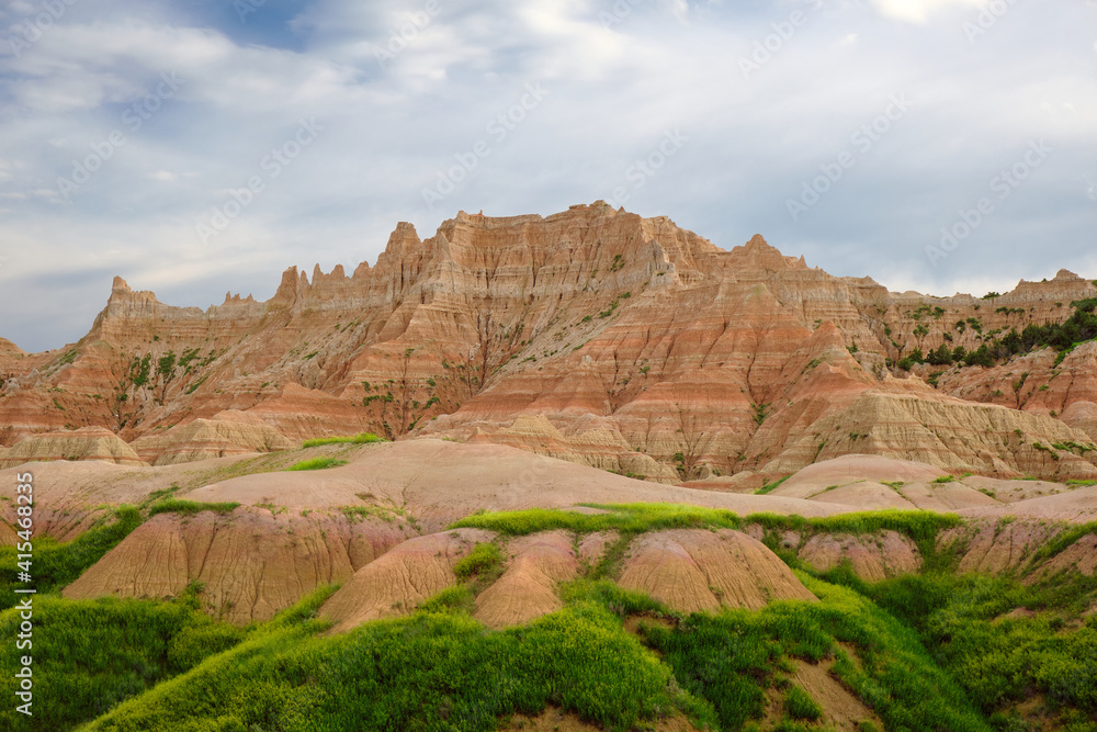 mountains in badlands national park. There is a lot of green grass growing in the foreground. The sky has a bit of blue, but is mostly cloudy