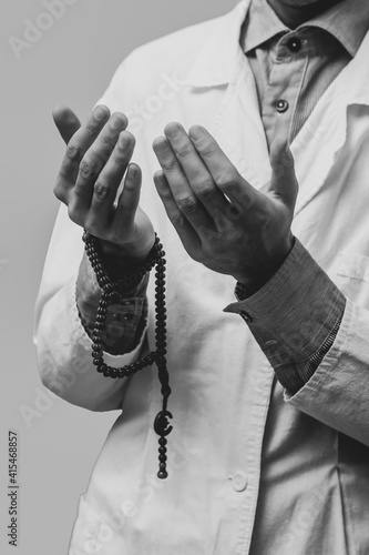 A young man in a white robe raised his hands up and prayed with a Muslim rosary. Medical gloves. Doctor praying. A Muslim makes dua. Black and white photo.