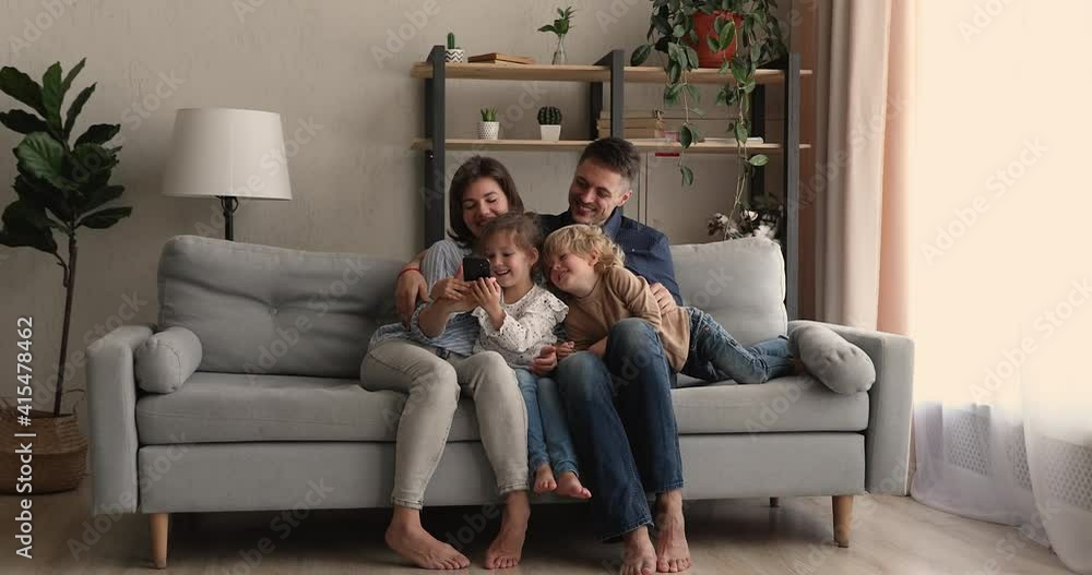 Full length happy young parents resting on sofa with little adorable children siblings, showing entertaining mobile applications, making funny photo or recording video on smartphone, tech addiction. Full length happy young parents resting on sofa with little adorable children siblings, showing entertaining mobile applications, making funny photo or recording video on smartphone, tech addiction.