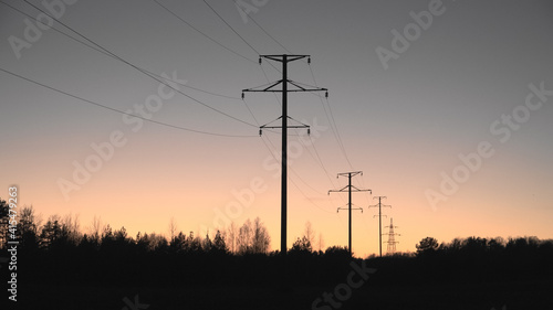 High voltage line on a sunset sky background. The black silhouettes of electric poles in a perspective view. Countryside environment with sundown heaven in the late evening to the nightfall time. © Uldis