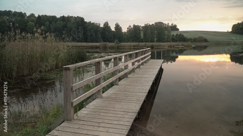 A wooden footbridge by the lake in wild nature landscape. A recreation area scenic view in the evening at sunset time. The calm surface of the water reflects sunlight. Grassy lakeshore a scenic view.