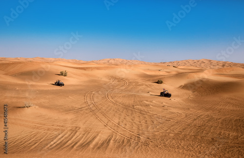 Beautiful Dubai desert landscape with plants and two riding quad buggy vehicles, sand with wheel tracks and blue sky
