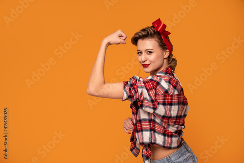 Studio shot of beautiful young woman with blond hair in pinup style clothes showing muscles to the camera, smiling.