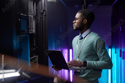 Side view portrait of African American data engineer holding laptop while working with supercomputer in server room lit by blue light, copy space photo