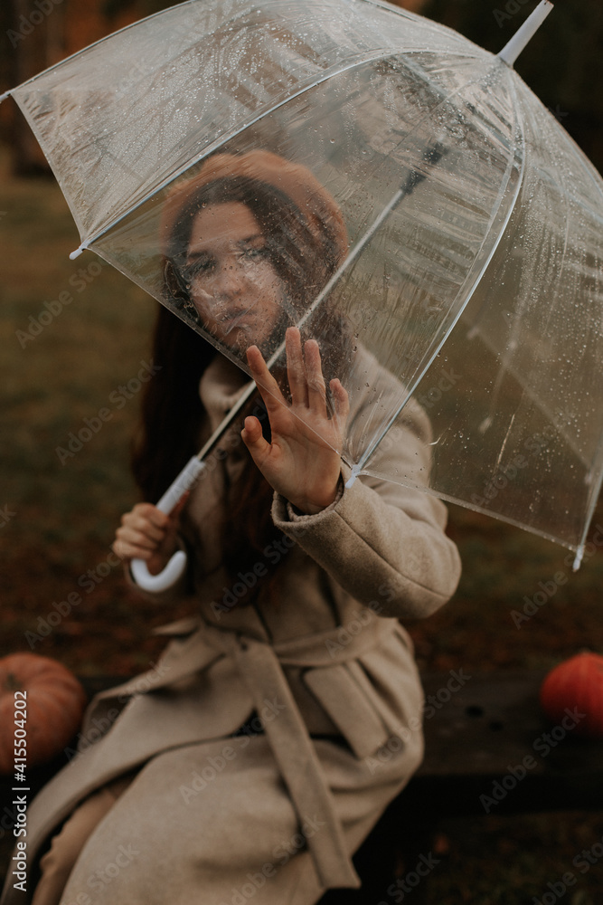 Fototapeta premium The girl sits on a bench, her hand touches the edge of the umbrella and looks through the transparent umbrella.