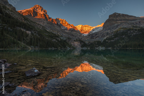 Sunset during fall in the mountains with snow, warm orange light on the peak, reflections on lake with calm clear water, trees and rocks, Lake O'Hara, Yoho National Park, British Columbia, Canada