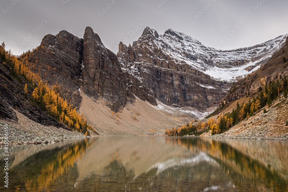 Mountain peaks with snow during fall, rocks, yellow and green larches along a lake with clear reflections under a gray sky. Lake Louis, Lake Agnes, Banff National Park, Alberta, Canada.