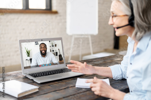 Senior gray-haired female therapist in headset looking at screen, talking to a young black man, having virtual consultation with client, online video session or a meeting via laptop from home office photo