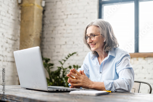 Senior businesswoman sitting at the desk, looking at webcam and talking with client online or watching tutorial, elder female office worker or therapist having appointment, virtual meeting via laptop photo