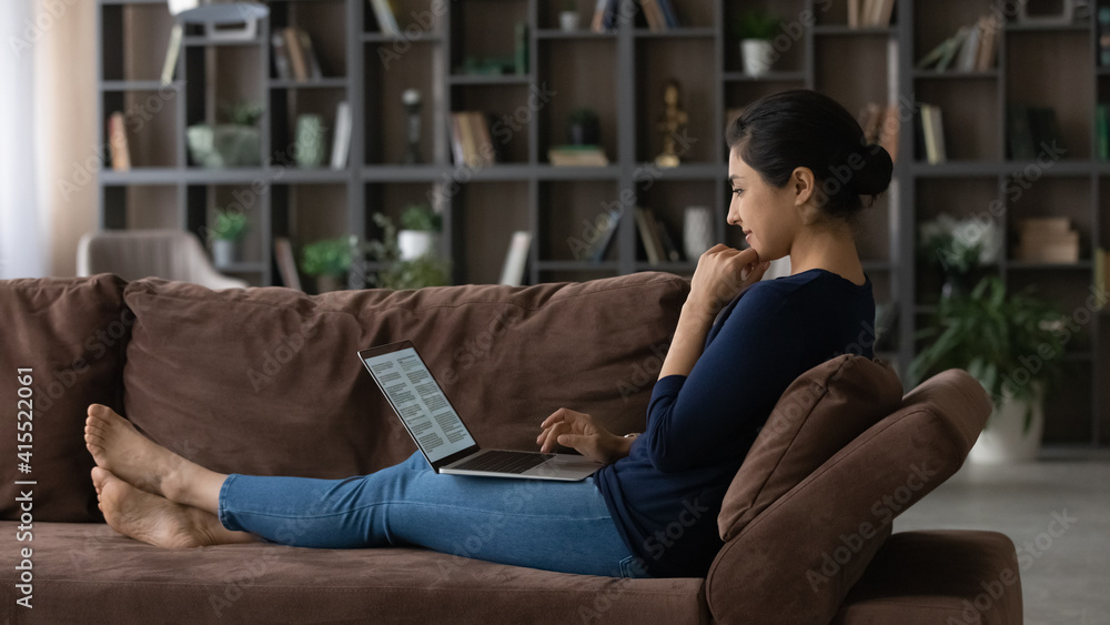 Millennial Indian woman sit relax on couch in living room work online. Young mixed race female rest on sofa at home study distant on laptop, prepare project or report on device.