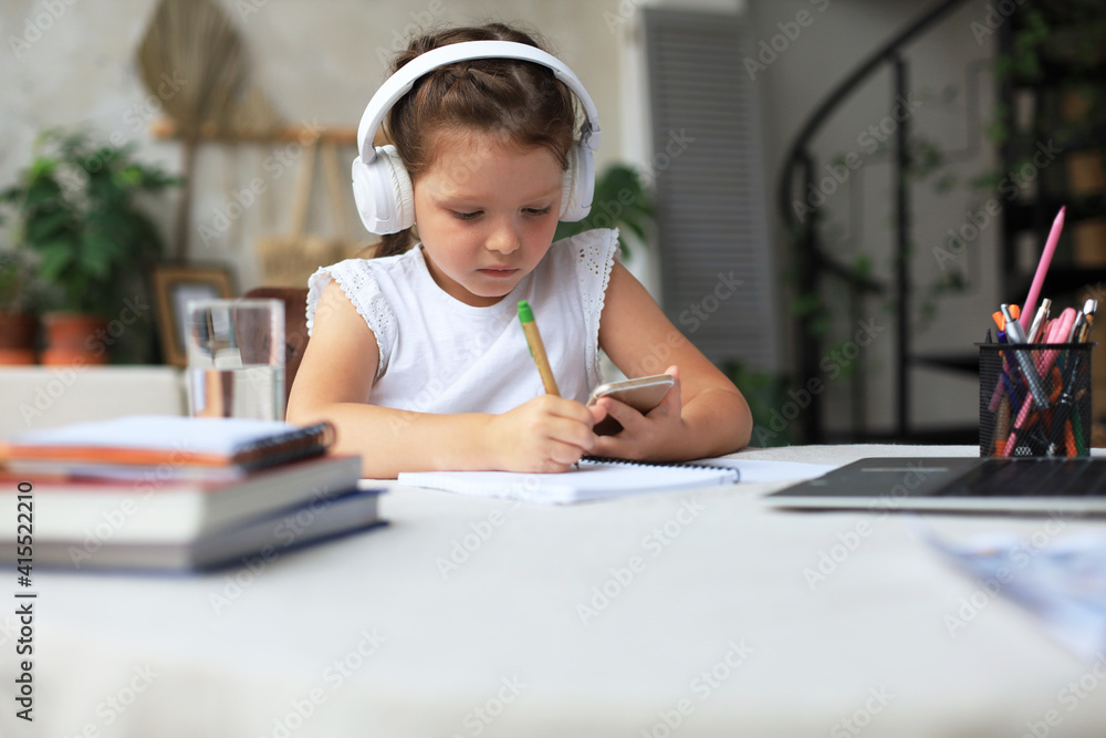 Smart little girl in headphones do homework online class on smartphone, small child in earphones study on Internet, have web conference or lesson during quarantine. Smart little girl in headphones do homework online class on smartphone, small child in earphones study on Internet, have web conference or lesson during quarantine.