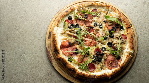 Ready-made round Italian pizza with cheese, salami and olives on a round wooden board on a gray background.