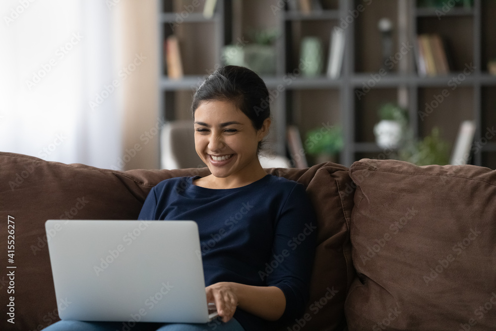 Naklejka premium Smiling millennial Indian woman relax on couch in living room look at laptop screen talk on video call. Happy young mixed race female use computer watch movie, have webcam online virtual conference.