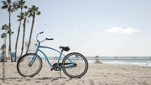 Blue bicycle, cruiser bike by sandy ocean beach, pacific coast, Oceanside pier California USA. Summertime vacations, sea shore. Vintage cycle, palms, sky, lifeguard tower watchtower hut, car truck.