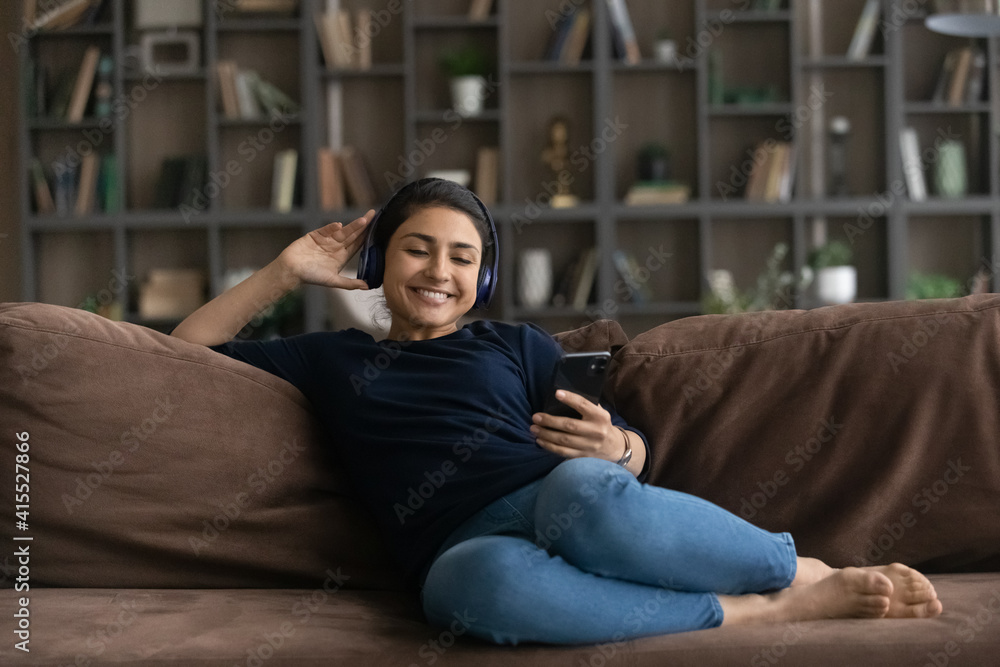 Happy young Indian woman relax on sofa in living room wear headphones listen to music on smartphone. Smiling millennial ethnic female rest at home enjoy good quality sound in earphones use cellphone. Happy young Indian woman relax on sofa in living room wear headphones listen to music on smartphone. Smiling millennial ethnic female rest at home enjoy good quality sound in earphones use cellphone.
