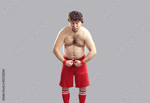 Fototapeta Funny topless strong plump athlete in red gym shorts showing his chubby body standing isolated on gray background