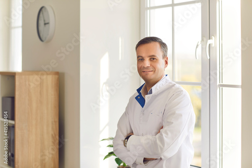 Smiling young man in white lab coat standing arms folded by window in laboratory or hospital office. Portrait of friendly confident trustworthy doctor, medical worker or scientist at work