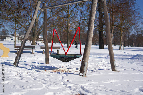Large swing on a playground in the park, trees and building in the background. red dominates. Blue sky. Winter with snow in germany.