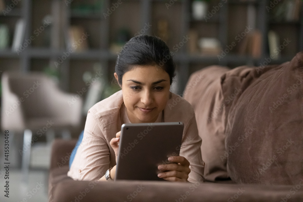 Millennial Indian woman lying on couch at home look at tablet screen text or message online. Happy young mixed race female relax in living room use pad device, browse internet. Communication concept. Millennial Indian woman lying on couch at home look at tablet screen text or message online. Happy young mixed race female relax in living room use pad device, browse internet. Communication concept.