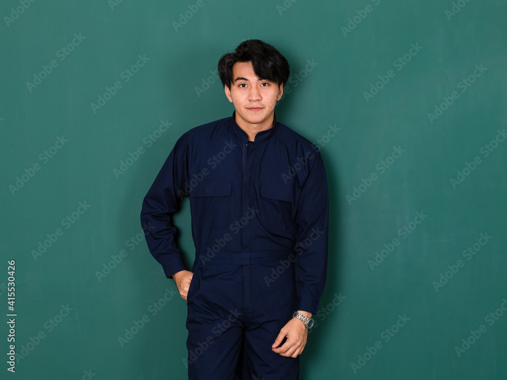 portrait of young handsome Asian man wearing a blue mechanic jumpsuit standing confidently and right hand in a pocket looking to a camera over green background