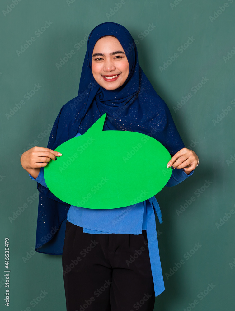 confident and happy woman concept. Pretty attractive Muslim lady dressed beautifully wearing blue hijab standing and holding green blank speech bubble over green background confident and happy woman concept. Pretty attractive Muslim lady dressed beautifully wearing blue hijab standing and holding green blank speech bubble over green background