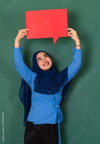 confident and happy woman concept. Pretty attractive Muslim lady dressed beautifully wearing blue hijab standing and holding a red blank speech bubble over green background © Bangkok Click Studio