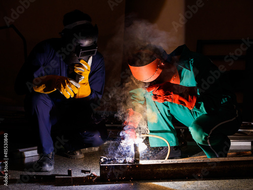 two engineers working in the dark at nighttime. Mechanics wearing a mechanic coveralls work together to weld the metal rod, making nice spark, bokeh, and smoke