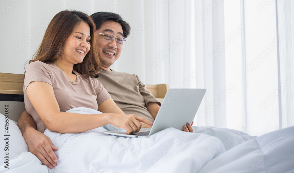 happy middle age Asian couple dressed casually sitting relaxed on a bed at home enjoy looking at a laptop computer. Warmth, delightful family concept happy middle age Asian couple dressed casually sitting relaxed on a bed at home enjoy looking at a laptop computer. Warmth, delightful family concept