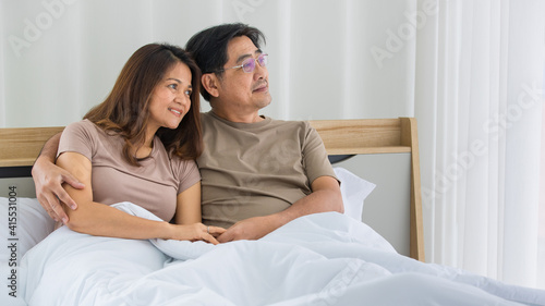 portrait of happy Asian middle-aged couple sitting on a bed, husband put his hand over the shoulder of the wife smiling to a camera in a bedroom. Warmth family concept