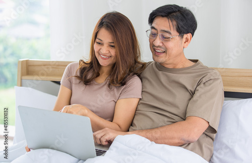 happy middle age Asian couple dressed casually sitting relaxed on a bed at home enjoy looking at a laptop computer. Warmth, delightful family concept