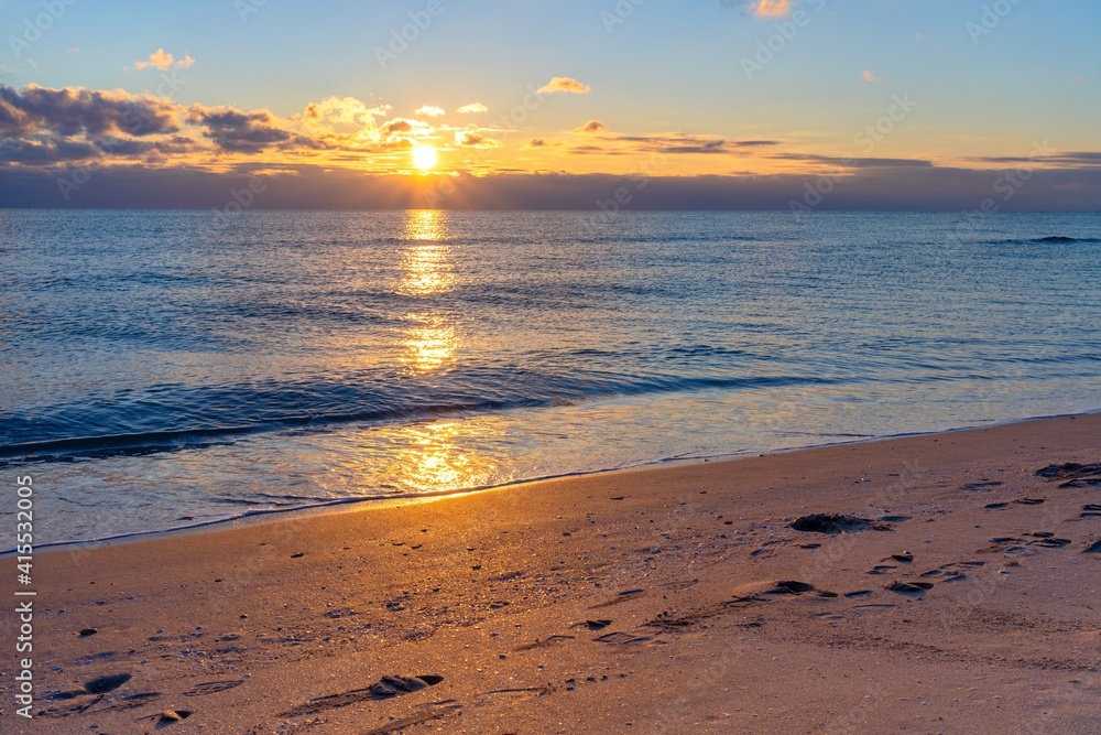 Golden rays of the setting sun are reflected on the water surface of the sea and the sandy shore on which traces of people are visible