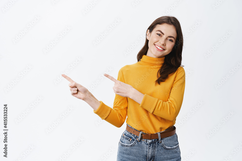 Cheerful smiling woman pointing fingers at copy space and staring at camera with pleasant and friendly expression, standing against white background. Advertising concept Cheerful smiling woman pointing fingers at copy space and staring at camera with pleasant and friendly expression, standing against white background. Advertising concept
