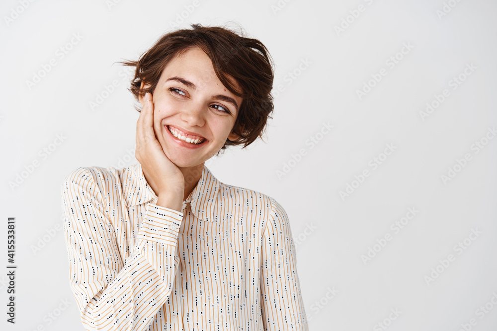 Portrait of cute brunette woman in blouse, touching well nourished facial skin with no makeup, natural beauty, looking left and smiling, standing over white background Portrait of cute brunette woman in blouse, touching well nourished facial skin with no makeup, natural beauty, looking left and smiling, standing over white background
