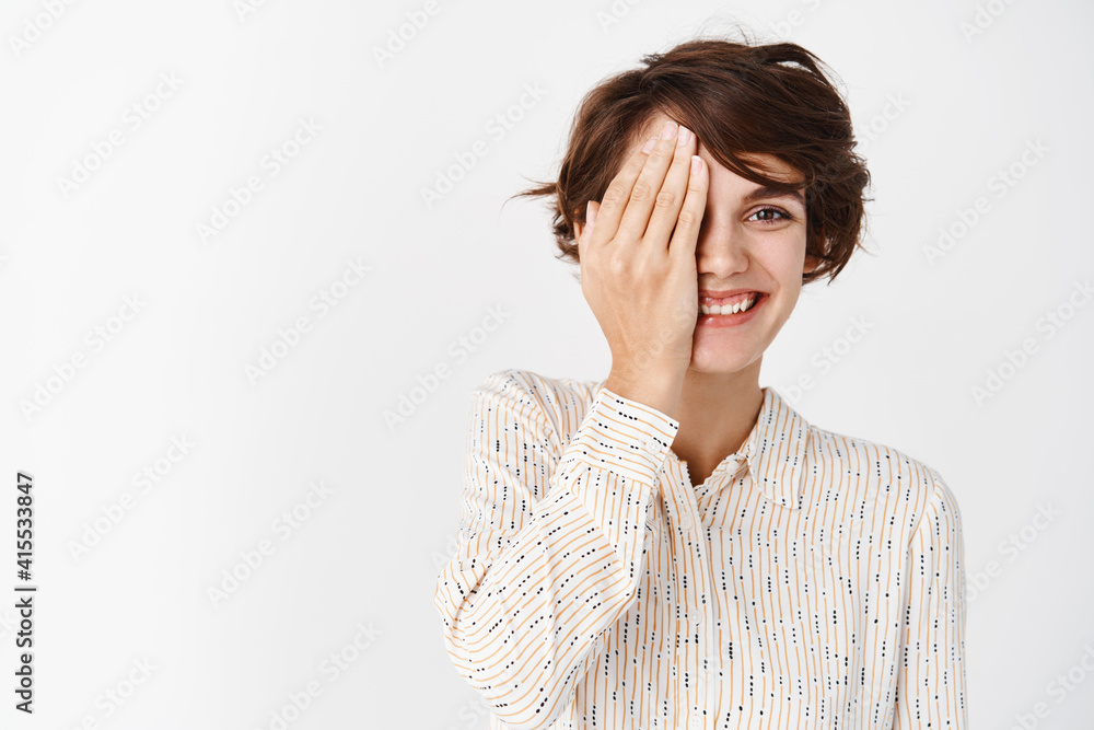 Portrait of smiling girl with short hair and natural beauty without makeup, cover half of face with palm and looking happy, standing against white background Portrait of smiling girl with short hair and natural beauty without makeup, cover half of face with palm and looking happy, standing against white background