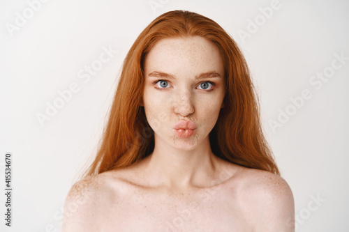 Close-up of beautiful young woman with long ginger hair. Redhead female with pale perfect skin, standing no makeup and pucker lips in kiss, looking with blue eyes at camera