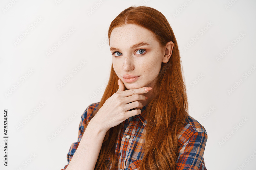Close-up of young female student with red natural hair, pale skin and blue eyes, looking thoughtful at camera, making choice at stare at camera, standing over white background Close-up of young female student with red natural hair, pale skin and blue eyes, looking thoughtful at camera, making choice at stare at camera, standing over white background