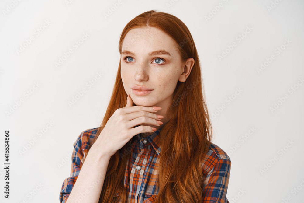Young redhead woman looking at upper left corner logo and thinking, making decision, touching chin with thoughtful face, standing over white background Young redhead woman looking at upper left corner logo and thinking, making decision, touching chin with thoughtful face, standing over white background
