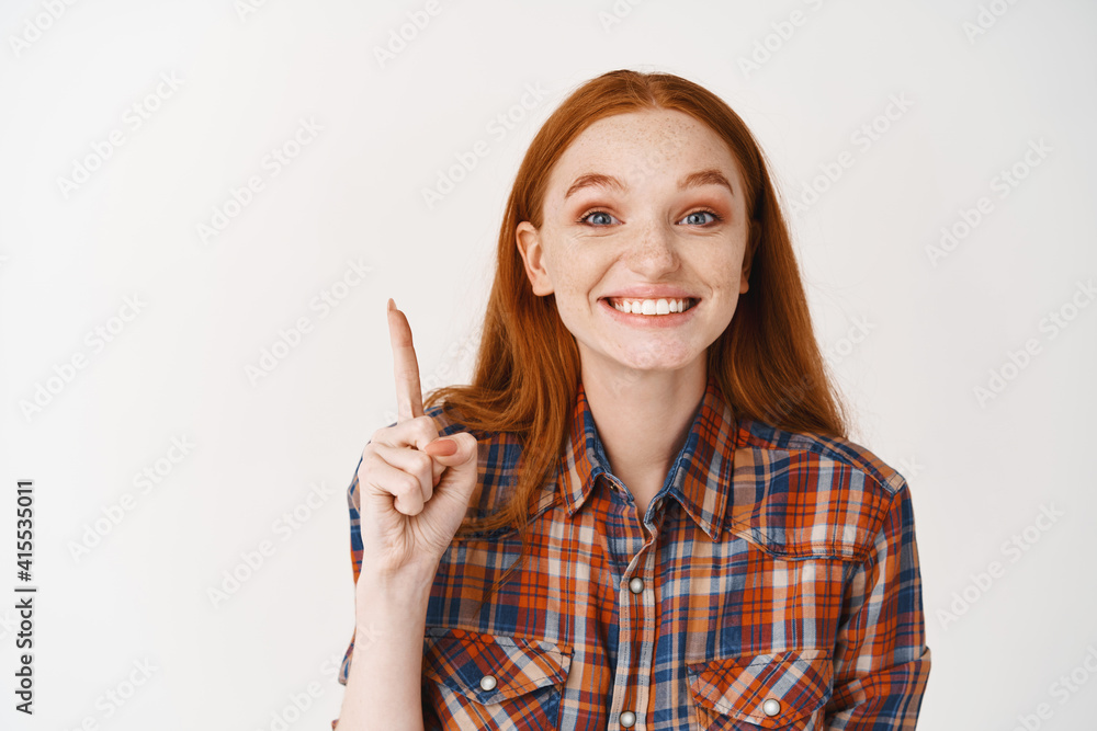 Cheerful redhead girl pointing finger up and smiling with white teeth, showing advertisement, standing over white background Cheerful redhead girl pointing finger up and smiling with white teeth, showing advertisement, standing over white background