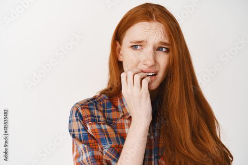 Close-up of scared and nervous woman with red hair  biting fingernails and frowning insecure  looking left worried  standing over white background