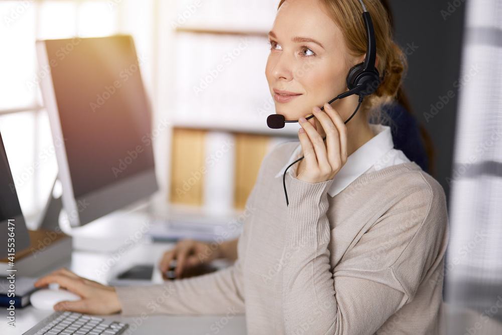 Businesswoman talking by headset while sitting in sunny office. Call center and diverse people group in business