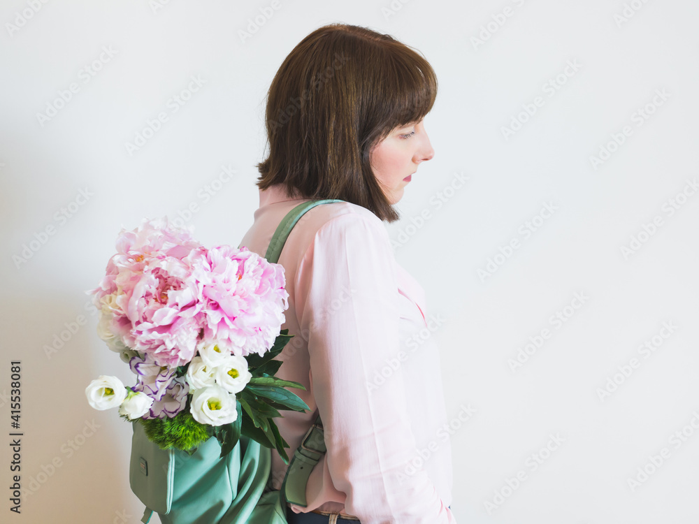 Young woman wearing pink shirt and backpack with pastel peony bouquet on gray wall background. Lifestyle spring concept. Mothers day gift Young woman wearing pink shirt and backpack with pastel peony bouquet on gray wall background. Lifestyle spring concept. Mothers day gift